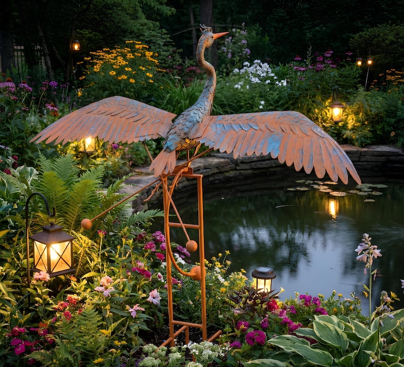 A large metal sculpture of a heron stands on a metal stand in a garden, surrounded by colorful flowers and plants. The heron's wings are spread wide, and it is perched on a ladder, creating a whimsical and artistic scene.