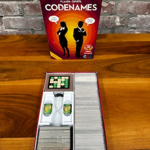 Pode incluir: A caixa do jogo de tabuleiro Codenames, com um design vermelho e laranja com duas silhuetas de pessoas. A tampa da caixa está aberta, revelando o tabuleiro de jogo, as cartas e um relógio de areia.