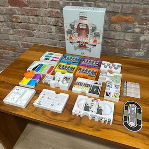 May include: The board game "Kanban EV" is displayed with its components on a wooden table. The box art shows a car design. The game includes coloured trays, cards, and tokens. The game appears to be a strategy or simulation game.
