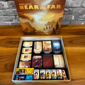 May include: An open box game organiser for the board game "Near and Far" by Ryan Laukat. The organiser has compartments for cards, dice, tokens, and player pieces. The box is made of cardboard and has a light blue and white colour scheme.