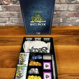 Puede incluir: Una caja azul con el texto "Isle of Skye: From Chieftain to King" y "Big Box" en ella. La caja está abierta y muestra el contenido del juego, incluyendo cartas, fichas y una bolsa de tela.