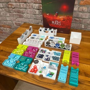 Op de afbeelding: Het bordspel "Mars" met een doos met een Marslandschap. Verschillende spelonderdelen in kleuren als blauw, groen en roze zijn gerangschikt op een houten tafel. Het woord "Mars" is zichtbaar.