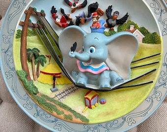 May include: A blue and gray ceramic plate with a 3D image of Dumbo the elephant riding on a swing with crows flying around him. The plate has a blue and white border with a decorative design.