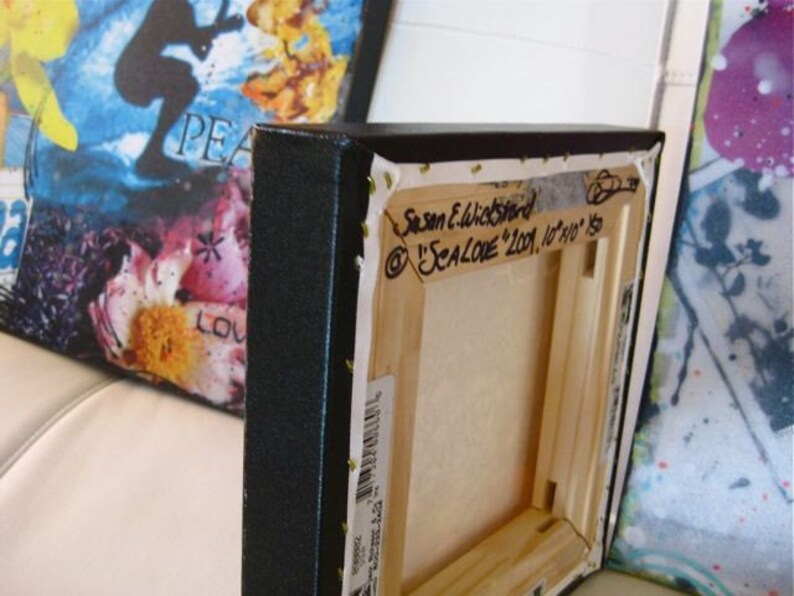 Puede incluir: Primer plano de la parte posterior de una pintura sobre lienzo enmarcada. El lienzo es negro con un marco de madera. La firma del artista y el t&iacute;tulo est&aacute;n escritos con marcador negro en la parte posterior del lienzo. El t&iacute;tulo es "SEALOVE 2001, 10"x10" "