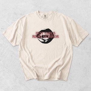 May include: A cream-coloured short-sleeve t-shirt featuring a graphic of black lips with sharp fangs. The text "HOT GIRLS READ HORROR" is printed in a red, slightly blurred font across the centre of the lips. The shirt has a crew neckline.