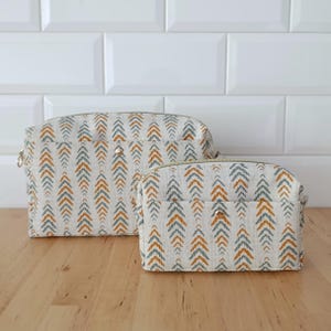 Peut inclure: Deux trousses rectangulaires zippées en tissu beige clair avec un motif chevron répétitif dans des tons bleu sourd et jaune moutarde. Chaque trousse comporte un bouton-pression métallique centré sur la poche extérieure avant.
