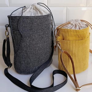 May include: Two drawstring bucket bags. One is black with a textured pattern and a black strap. The other is yellow corduroy with a brown leather strap. Both have patterned fabric lining the top.