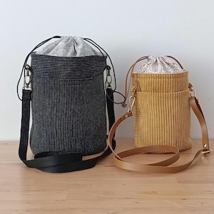 Puede incluir: Dos bolsos tipo cubo cilíndricos con cierre de cordón. El bolso más grande es negro con un patrón de cuadrícula texturizado y forro gris con estampado de conchas. El más pequeño es de pana color mostaza con forro floral y correa color canela.