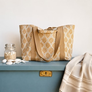 May include: A large tote bag with a leaf pattern in shades of gold and white. The bag has sturdy tan straps and a matching pocket. A jar of sand and seashells sits nearby, along with a folded beige and white striped towel. The scene is set on a blue surface.