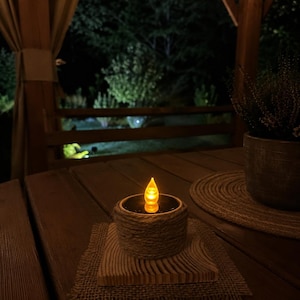 May include: A battery-operated candle with a flickering yellow flame sits on a wooden table. The candle is encased in a woven rope holder, placed on a wooden coaster and a burlap mat. The scene is set outdoors at night.