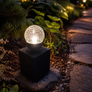 May include: A solar garden light featuring a spherical glass globe with a crackled texture, illuminated from within by a warm white light. The globe sits atop a solid black square base, providing a steady foundation for the outdoor fixture.