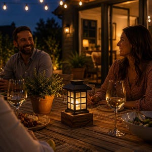 May include: A square, black, lantern-style lamp with a grid-patterned glass shade sits on a thick, natural-toned wooden base. The lamp emits a warm, soft glow from within, illuminating the center of a wooden outdoor dining table at night.