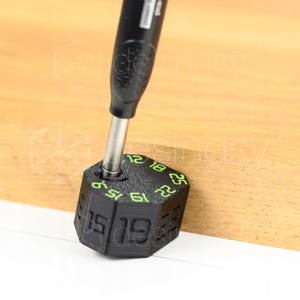 Può includere: Un utensile esagonale nero con superficie ruvida e numeri verde brillante stampati sulla parte superiore e sui lati. Una punta di cacciavite in metallo è inserita in una fessura sulla parte superiore. Sono visibili i numeri 9, 12, 15, 18, 19, 22 e 25.
