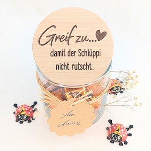 Könnte beinhalten: Ein klares Glasgefäß gefüllt mit orange verpackten Bonbons, verschlossen mit einem runden Holzdeckel, der den Text "Greif zu... damit der Schlüppi nicht rutscht." trägt. Ein gewelltes Etikett aus Kraftpapier ist mit "...für Anna" beschriftet.