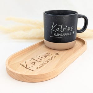 Könnte beinhalten: Eine schwarze, gesprenkelte Keramiktasse mit beigem Boden und Henkel, die auf einem ovalen Holztablett steht. Die Tasse und das Tablett tragen den Text "Katrins kleine Auszeit" in weißer Schrift. Ein Herzsymbol ist ebenfalls vorhanden.