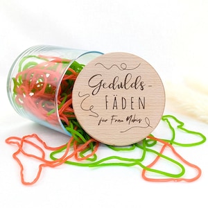 Könnte beinhalten: Ein durchsichtiges Glasgefäß gefüllt mit verwirrten neonorangen und hellgrünen Gummibändern, verschlossen mit einem runden Holzdeckel, der mit dem Text Gedulds-FÄDEN für Frau Nobis und dekorativen geschwungenen Linien graviert ist.