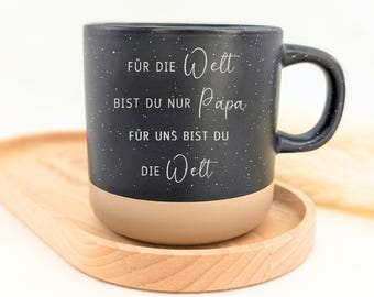 May include: A two-toned ceramic mug with a dark charcoal speckled upper section and a solid light beige base. The front features white text that reads: FÜR DIE Welt BIST DU NUR Papa FÜR UNS BIST DU DIE Welt.