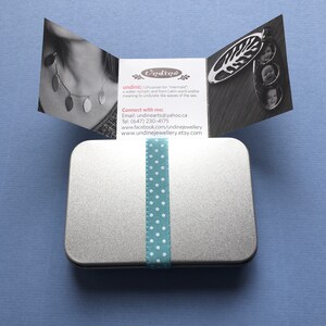 May include: A silver metal tin with a blue and white polka dot ribbon tied around it. The tin is open and a card with the word "Undine" is visible. The card explains that "undine" is Lithuanian for "mermaid" and that it is a water nymph. The card also includes contact information for the seller.