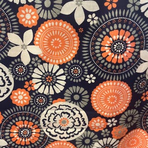 May include: Close-up of a fabric with a repeating floral pattern. The design features white, orange, and gray flowers and circular motifs on a dark navy blue background. The pattern is dense and detailed, creating a visually rich textile.