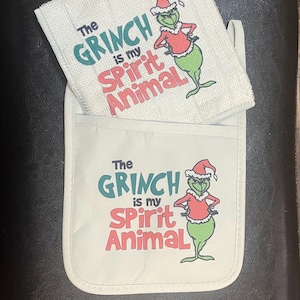 May include: A white oven mitt and matching towel set featuring the Grinch character. The text "The Grinch is my Spirit Animal" is printed in green and red. The Grinch is wearing a Santa hat and red shirt.