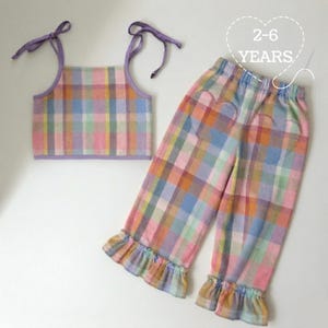 Könnte beinhalten: Ein zweiteiliges Kinder-Outfit in einem farbenfrohen Karomuster. Das Set besteht aus einem Crop-Top mit lila Bindeträgern und einer passenden Hose mit herzförmigen Details und Rüschenabschlüssen. Der Text "2-6 YEARS" ist ebenfalls sichtbar.