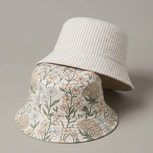 May include: A reversible bucket hat featuring two distinct sides. One side displays a cream-coloured fabric with a repeating pattern of small white daisies and light peach-coloured flowers with green stems. The other side has a beige and white striped pattern.