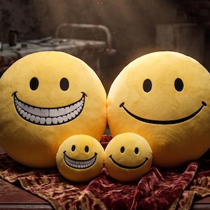 May include: Two large and two small round yellow plush pillows featuring black embroidered eyes and smiles. One large and one small pillow display a wide, toothy grin with visible white teeth, while the other two feature a simple curved line smile.
