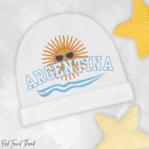 May include: White baby beanie featuring the word "ARGENTINA" in blue block letters, with a sun wearing sunglasses graphic. A blue wave design is below the text. Two yellow star-shaped toys are in the background.