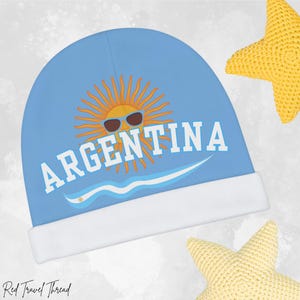 May include: A blue beanie with a white brim features a yellow sun with sunglasses and the word "ARGENTINA" in white letters. The sun is surrounded by rays and there is a blue wave below it.