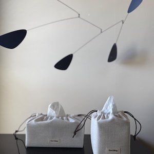 May include: Two light gray fabric tissue box covers with a drawstring closure and a small label that reads "from liking". A modern mobile with black teardrop shapes hangs in the background.