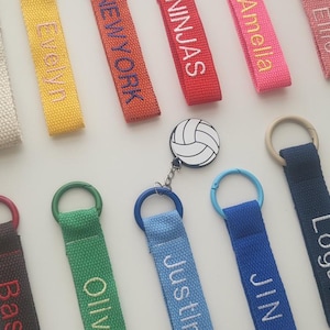 May include: A collection of colorful keychains with embroidered names and designs. The keychains are made of fabric and have metal rings. Some of the designs include a volleyball, a piano, and the words "Golfholics" and "JIN".
