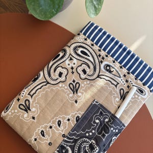 Op de afbeelding: Een gewatteerde stoffen hoes met een paisley patroon in beige, zwart en wit. De hoes heeft een marineblauwe en wit gestreepte rand en een klein vakje met een witte stylus. De hoes ligt op een bruin oppervlak.