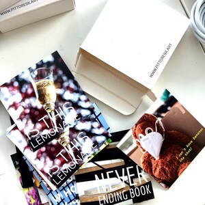 May include: A collection of square art prints and a white cardboard box with the text WWW.PITTORESK.ART. One print shows a champagne glass with the text IS THIS LEMONADE, another shows a brown plush toy wearing a mask with the text I can't have bananas like that!, and a third shows the text NEVER ENDING BOOK.