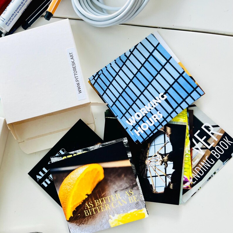 May include: A white cardboard box with the text WWW.PITTORESK.ART and a collection of square cards featuring various images and text, including the phrases WORKING HOURS and AS BITTER AS BITTER CAN BE, alongside a partial view of a book cover.