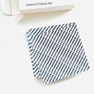May include: A square piece of white paper featuring a repeating black diagonal pattern of the text PITTORESK. Above the paper is a white box with the text WWW.PITTORESK.ART printed in black on the side. The items are arranged on a white surface.