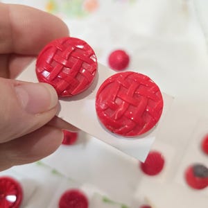 May include: Two circular red buttons featuring a raised, woven basket-weave texture on the surface. The buttons are mounted on a white card, displaying a glossy finish and a consistent, vibrant red colour throughout the intricate, crisscrossing pattern.