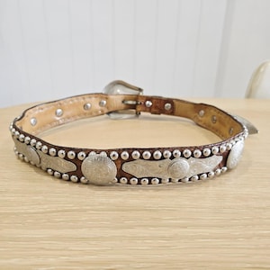 May include: A brown leather belt with silver decorative accents. The belt has a silver buckle and is decorated with silver studs and metal pieces. The belt is displayed on a light-coloured wooden surface.