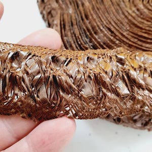May include: A roll of brown woven trim with a frayed edge. The trim is made of a thin, flexible material and has a delicate, intricate pattern.