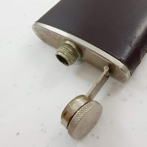 Può includere: Una fiaschetta in metallo argentato e marrone con un tappo a cerniera. La fiaschetta è rivestita in pelle e ha un tappo in metallo argentato.