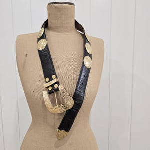 May include: Black belt with gold-coloured metal accents. The belt features a large, ornate buckle and decorative circular studs. The belt is displayed on a mannequin. The belt is made of a textured material.