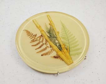 May include: A round, cream-colored coaster with embedded botanical elements. The coaster features dried ferns and bamboo stalks in shades of brown and yellow. The coaster is approximately 4 inches in diameter.