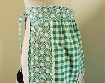 May include: A green and white gingham apron with a pink embroidered design. The apron has a pocket on the right side and ties at the waist.
