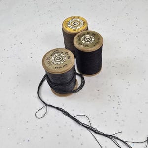 May include: Three vintage wooden spools of thread in shades of brown and black. The spools have labels with text and are arranged on a white surface. The thread is partially unwound from one spool.