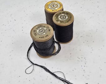 Könnte beinhalten: Drei Vintage-Holzspulen mit Faden in Braun- und Schwarztönen. Die Spulen haben Etiketten mit Text und sind auf einer weißen Oberfläche angeordnet. Der Faden ist teilweise von einer Spule abgewickelt.