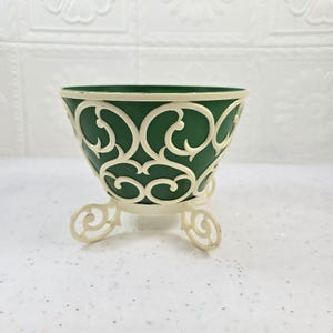 May include: A decorative green metal bowl with an off-white, ornate, openwork design. The bowl has three curved feet and a contrasting off-white rim. The bowl is likely for decorative purposes.