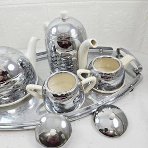 May include: A chrome tea set with a teapot, creamer, sugar bowl, and serving tray. The teapot and sugar bowl have white handles and spouts. The set is on a silver tray, reflecting the light.