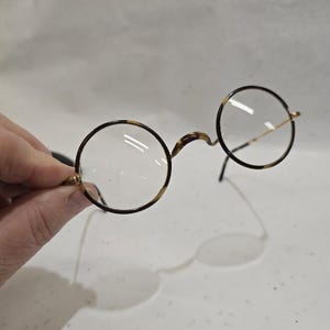 Puede incluir: Gafas redondas con montura con estampado de carey. Las lentes son transparentes y las monturas tienen un puente y varillas de metal dorado. Las gafas se sostienen sobre un fondo blanco.