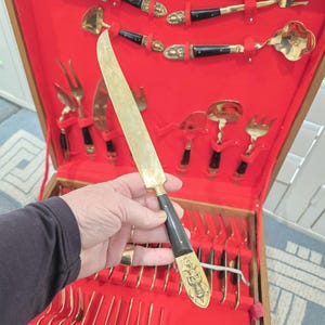 Könnte beinhalten: Ein Vintage-Tranchiermesser in Goldtönen mit schwarzem Griff, gehalten vor einer rot samtgefütterten Besteckkassette. Die Kassette enthält verschiedene goldfarbene Servierutensilien mit schwarzen Griffen und dekorativen Akzenten. Das Messer ist ca. 30 cm lang.