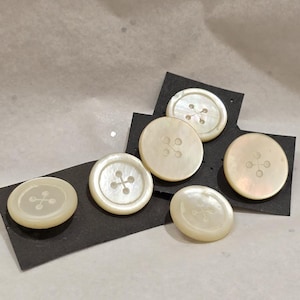 May include: A set of five round, off-white buttons with a pearlescent finish. Each button features a recessed centre with four small circular indentations connected by thin lines in a cross pattern, mounted on black rectangular display cards.
