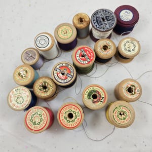 May include: A collection of vintage wooden spools of thread in various colours including red, blue, green, and white. Each spool features a circular paper label on top with brand names like J&P Coats, Clark & Co, and Dewhurst's, displaying text.